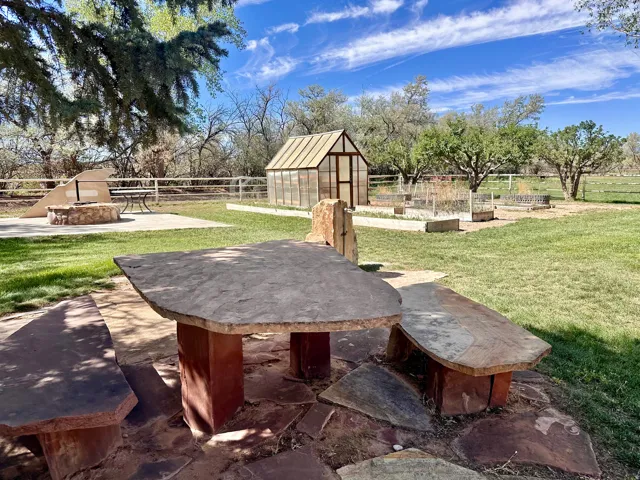 Expansive outdoor space featuring a stone table and benches, a built-in fire pit with surrounding concrete patio, a greenhouse structure, and raised garden beds