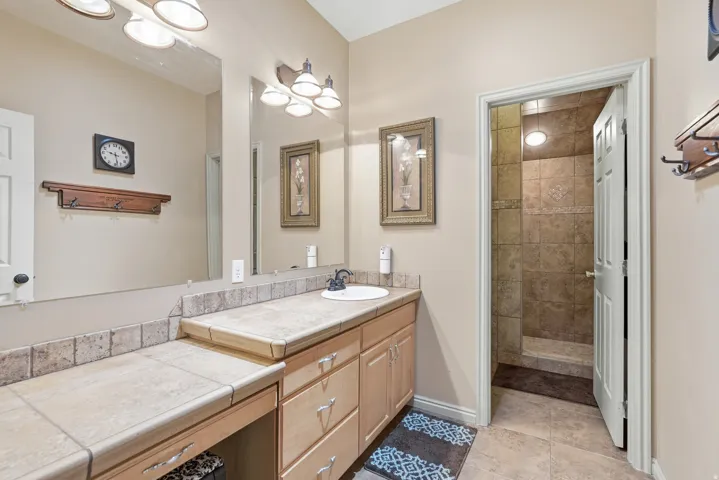 Vanity with integrated sink, tile countertop, wood cabinetry, and an adjacent tiled shower with a built-in bench