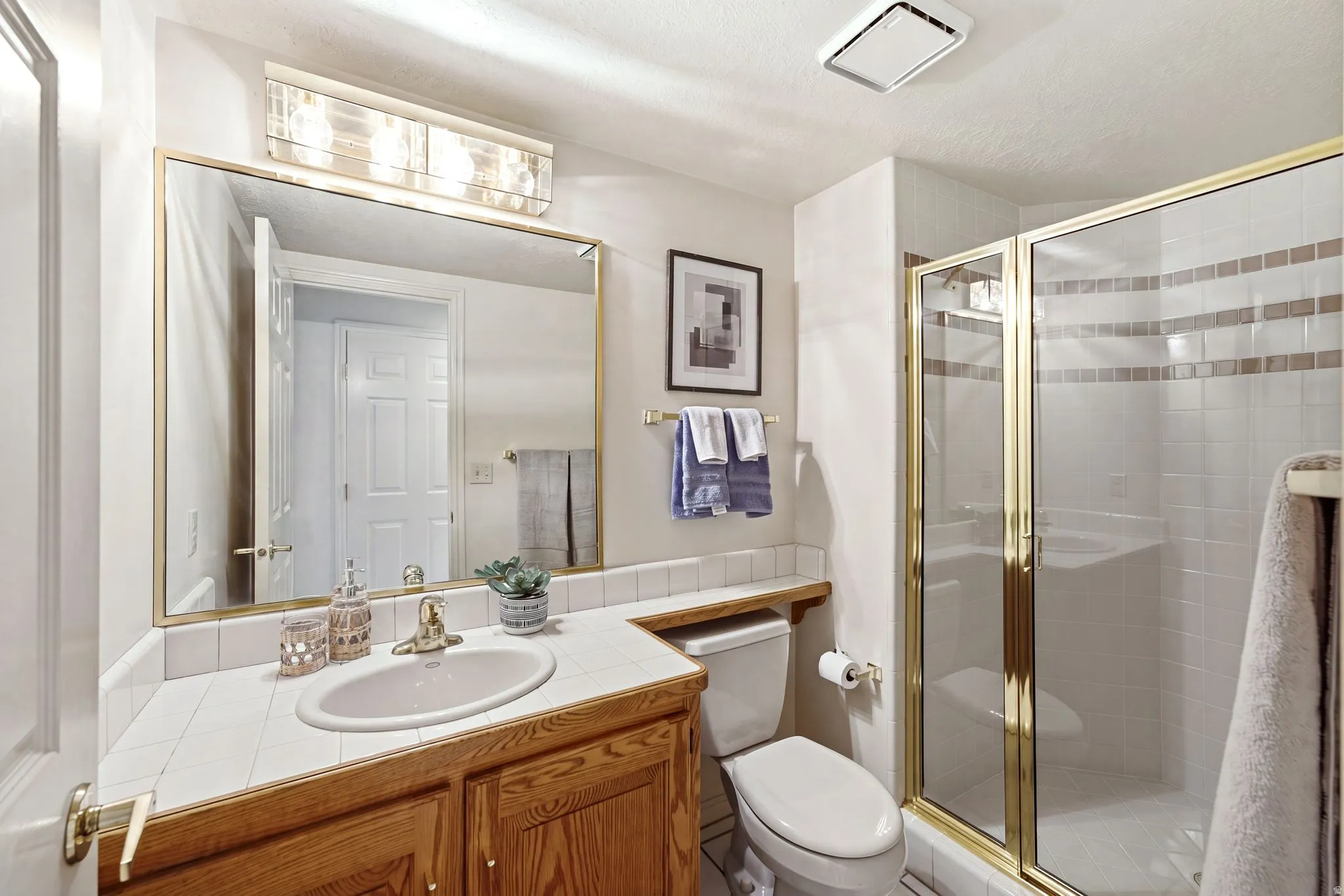 Bathroom featuring a wood-finish vanity with an integrated sink, white tiled countertop, a framed mirror, and a standing shower with a glass enclosure