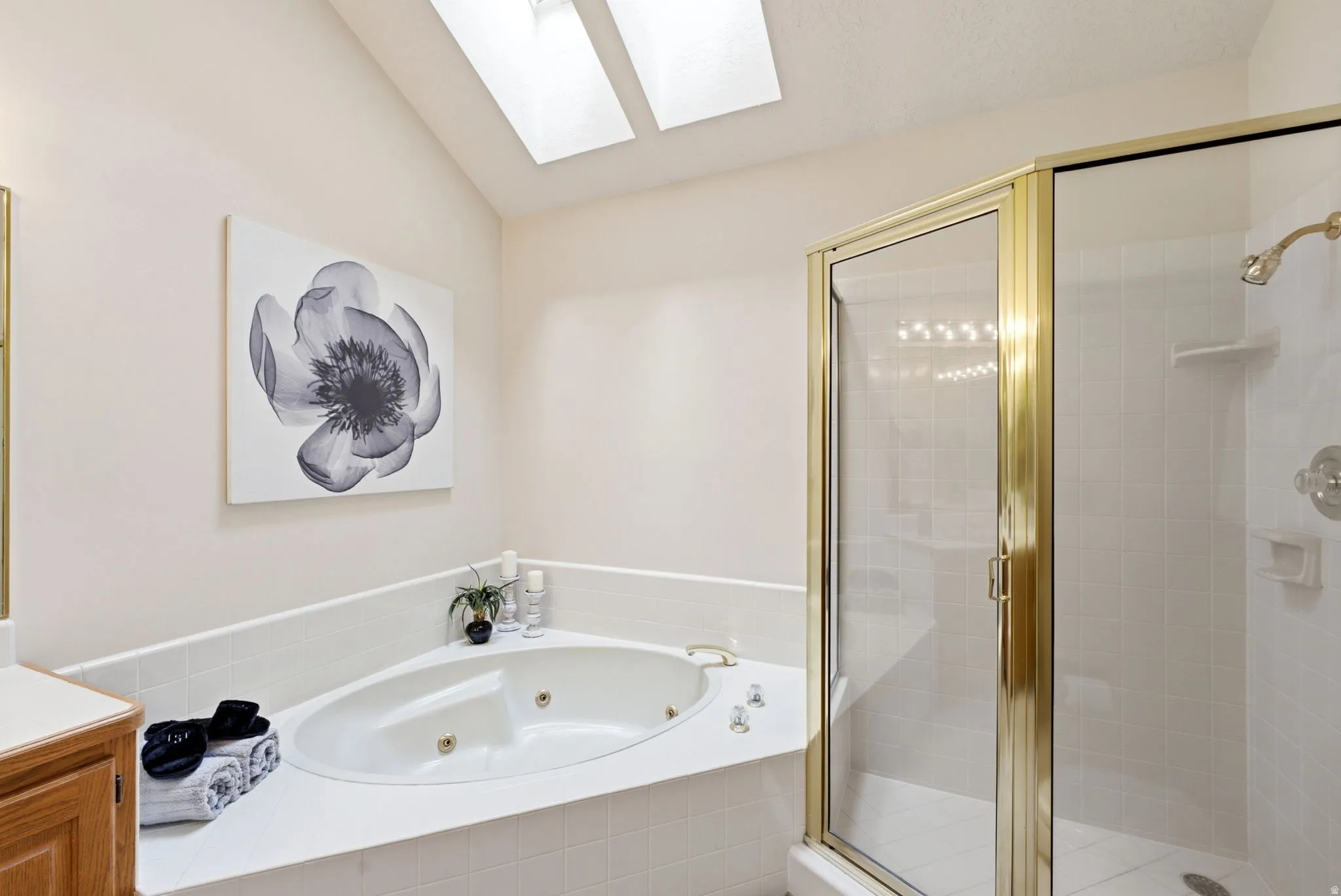 Soaking tub with jets, glass-enclosed shower with gold-tone framing, two ceiling skylights, and a tiled tub surround