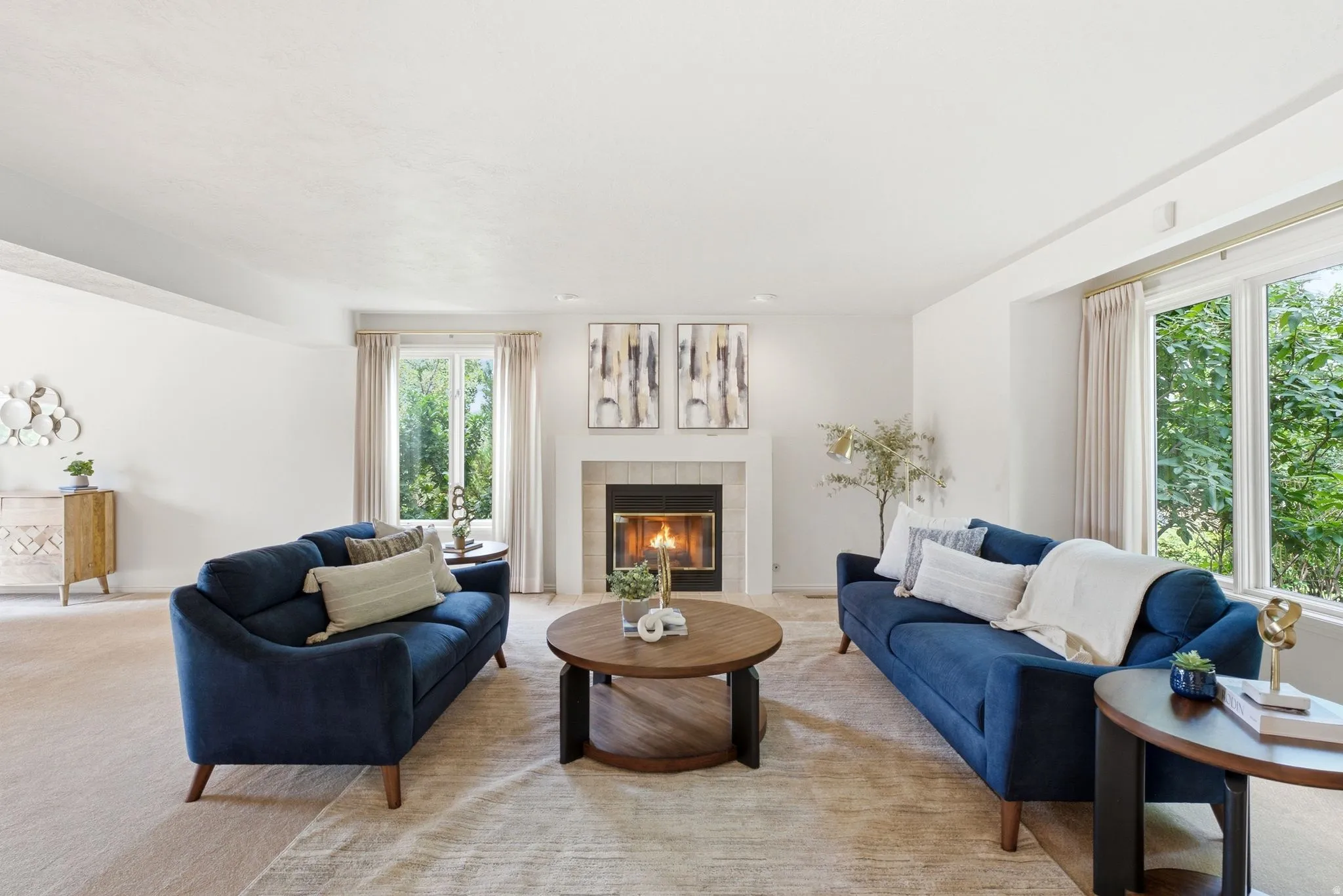 Spacious living area featuring a central fireplace with a tile surround and white mantel, large windows with exterior foliage, white walls, and light-toned carpeting