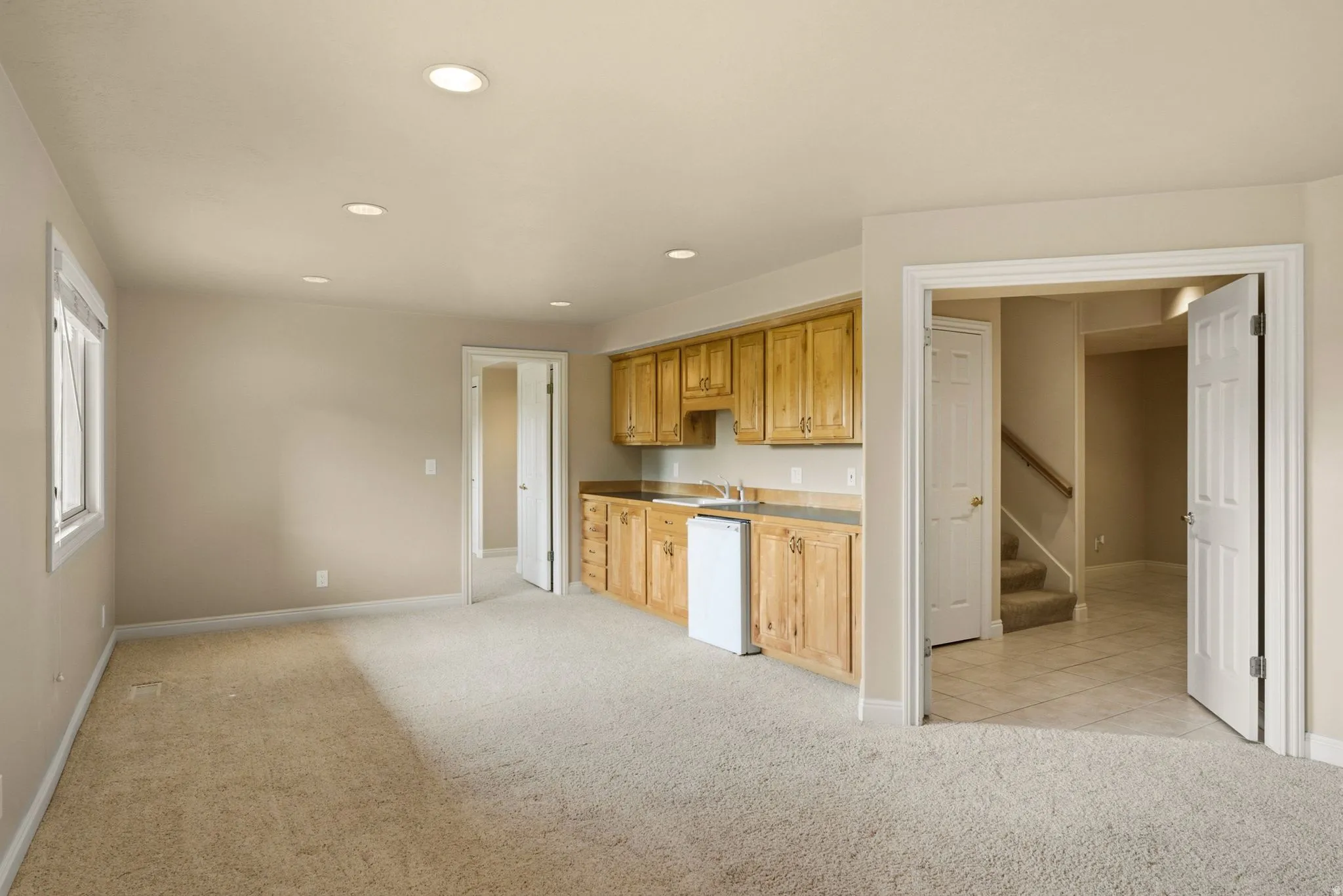 Recessed lighting illuminates the open area featuring carpeted flooring and a built-in wet bar with wood cabinetry and a compact refrigerator