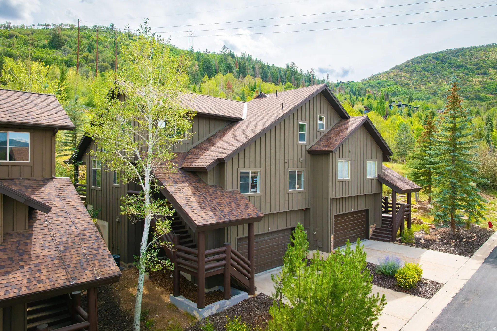 Mountain-style multi-level residence featuring brown vertical siding, multiple pitched roofs with asphalt shingles, attached garages with wood-finish doors, and mature evergreen trees