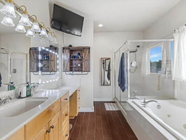 Primary bathroom featuring a dual vanity with light-colored countertops, a glass-enclosed shower, a jetted soaking tub and wood-finish flooring