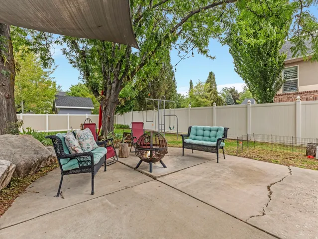 Spacious concrete patio featuring a shade sail, mature trees, a built-in fire pit, and a privacy fence