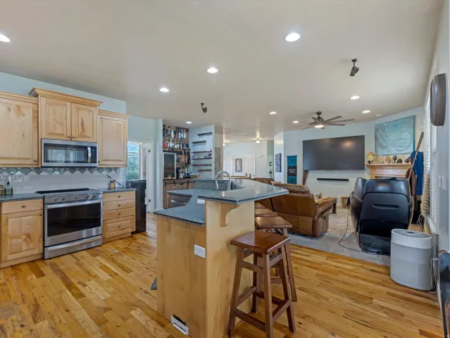 Open-concept kitchen featuring wood-finish flooring, light wood cabinetry, stainless steel appliances, a tiled backsplash, and an island with a breakfast bar