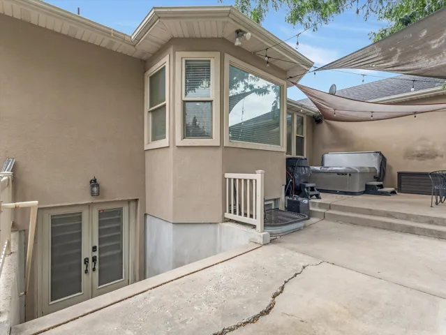 Lower-level exterior entrance featuring double doors with glass inserts, bay window with blinds, stucco siding, and a concrete patio with shade sails