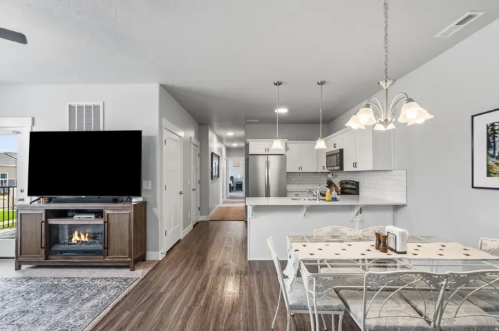 Open-concept living area featuring wood-finish flooring, a contemporary entertainment console with an integrated electric fireplace, a modern kitchen with white cabinetry and stainless steel appliances, a subway tile backsplash, and a breakfast bar