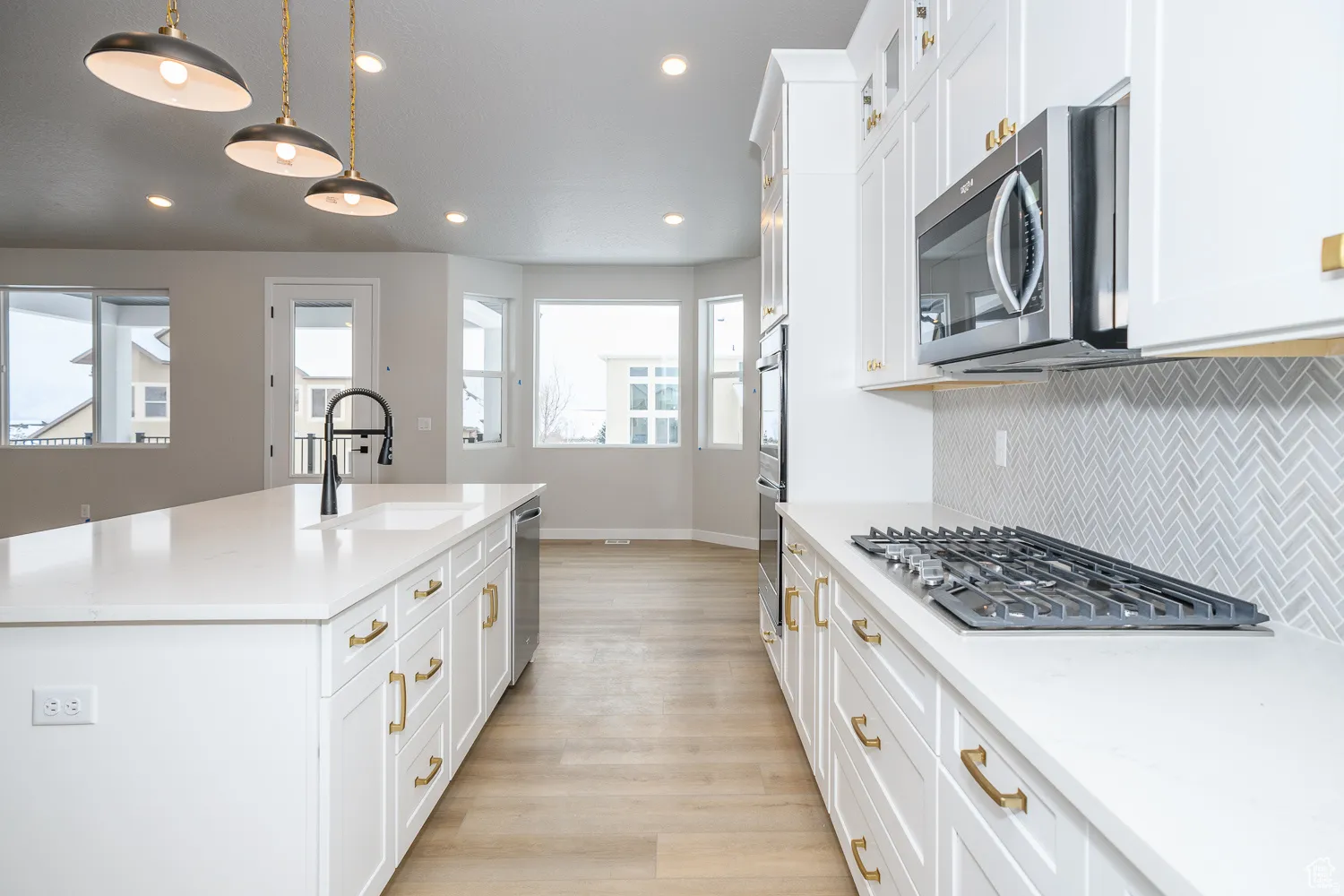 Spacious kitchen featuring an extended island with a built-in sink, white shaker cabinetry with gold-tone hardware, a tiled herringbone backsplash, and a gas cooktop with an overhead microwave