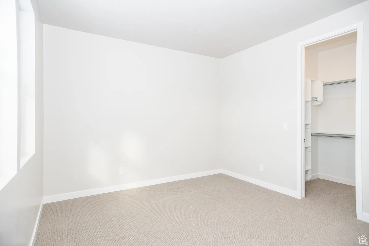 Carpeted room with white walls, baseboards, and ceiling, featuring a window and an open doorway revealing a closet with shelving and hanging rods