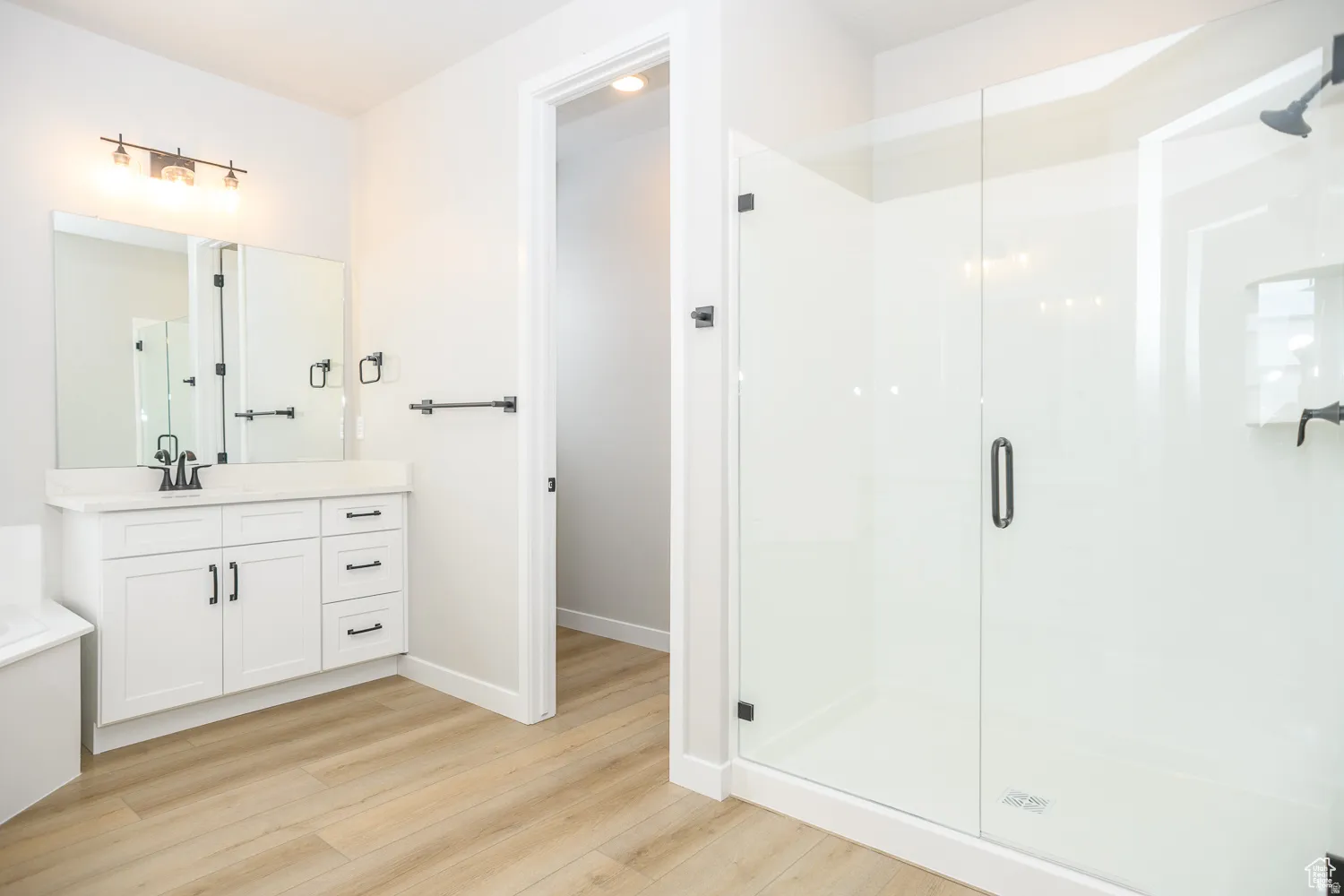 Bathroom featuring a glass-enclosed shower, white vanity with integrated sink, matte black fixtures, wood-finish flooring, and white walls