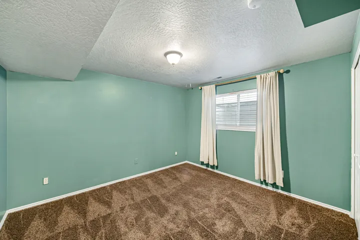 Interior room featuring a window with white blinds and drapes, textured ceiling, brown patterned carpet, and light green walls with white trim