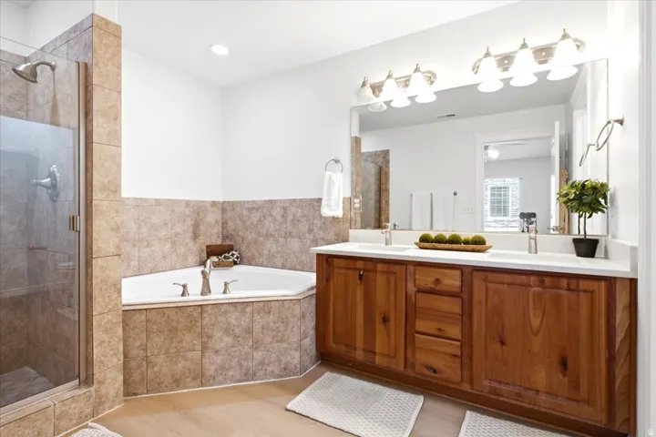 Bathroom featuring a corner soaking tub, a glass-enclosed shower, a double vanity with two sinks, wood-finish cabinetry, and a large wall mirror with overhead lighting