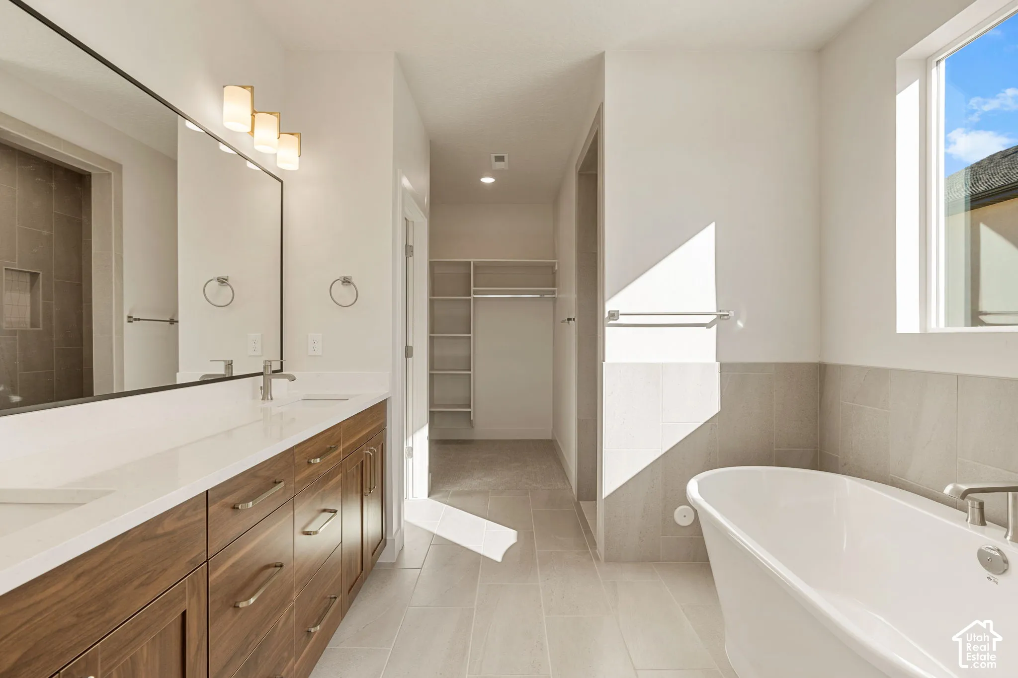 Full-width vanity with wood-finish cabinetry and white countertop, freestanding soaking tub, ceramic tile flooring, and walk-in closet with built-in shelving