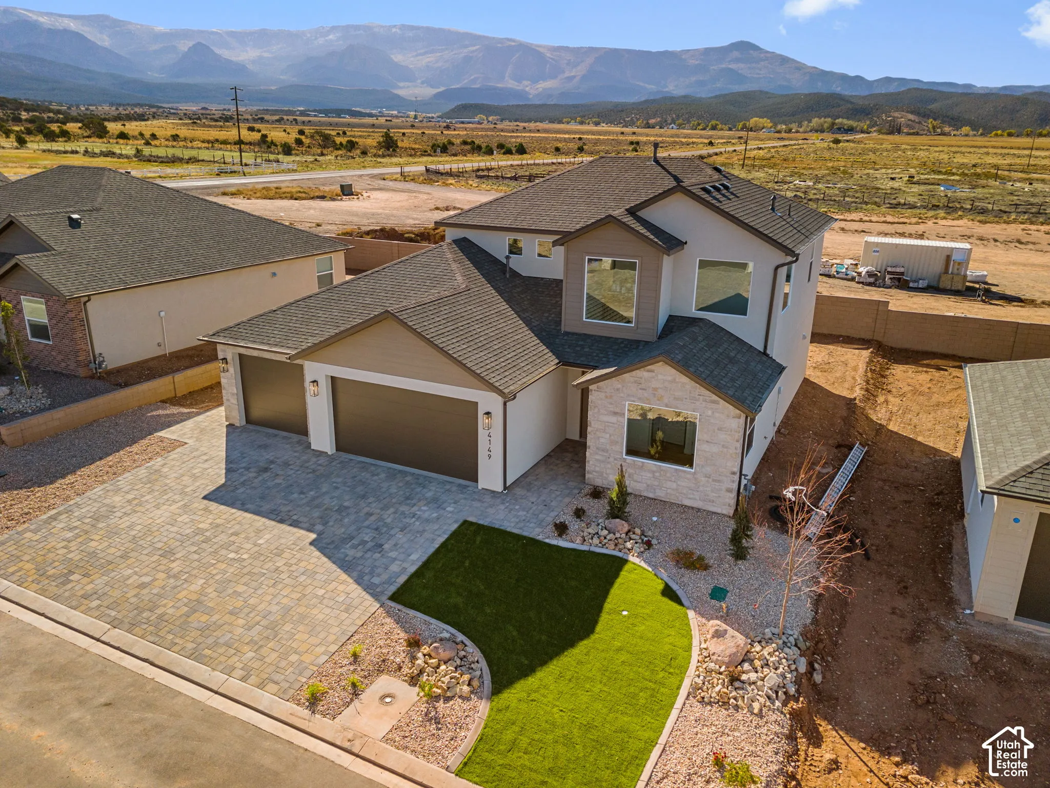 Two-story exterior featuring stone veneer accents, multi-tonal paver driveway, three-car garage, artificial turf lawn, and mountain backdrop