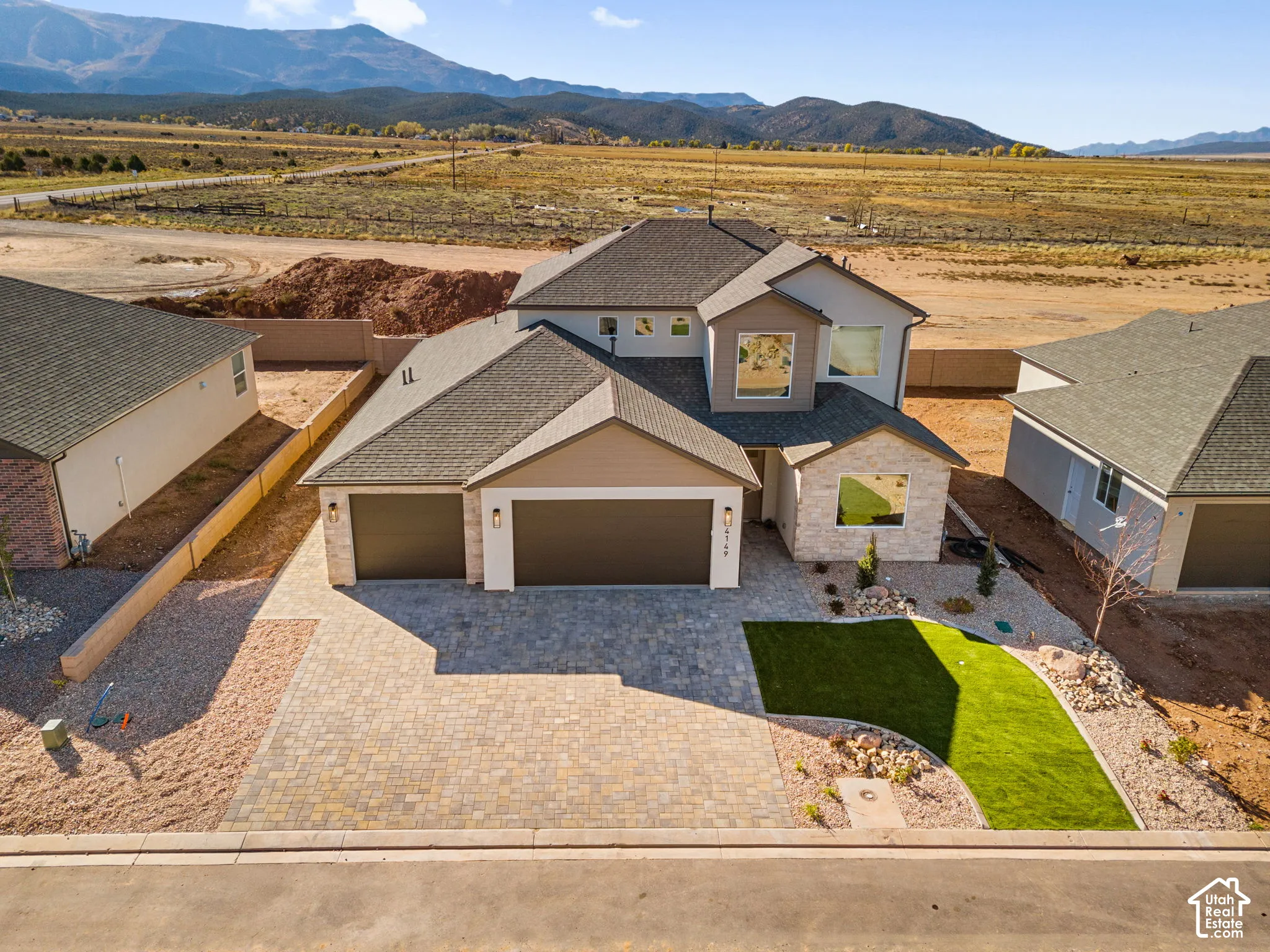 Two-story residence featuring a stone and siding exterior, multi-car garage, paver driveway, artificial turf lawn, and prominent mountain backdrop