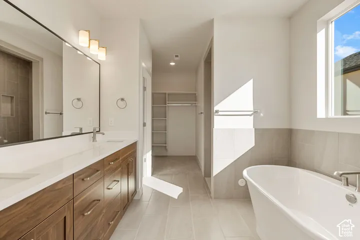 Full-width vanity with wood-finish cabinetry and white countertop, freestanding soaking tub, ceramic tile flooring, and walk-in closet with built-in shelving