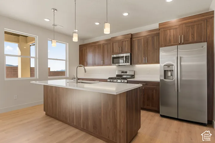 Kitchen featuring wood-finish cabinetry, quartz countertops, a central island with a built-in sink, stainless steel appliances, and recessed lighting
