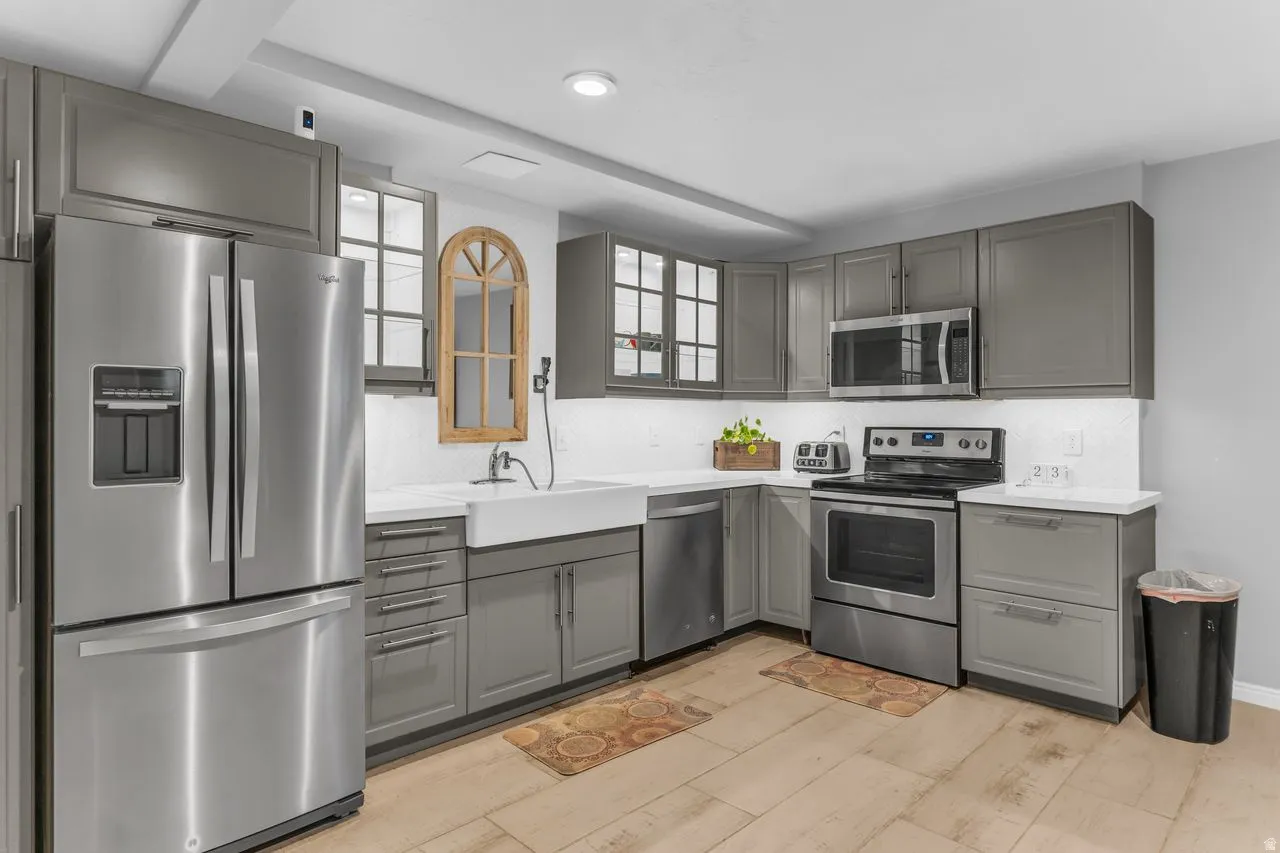 Basement kitchen featuring stainless steel appliances, a farmhouse-style sink, grey shaker cabinetry, white countertops, and wood-finish flooring