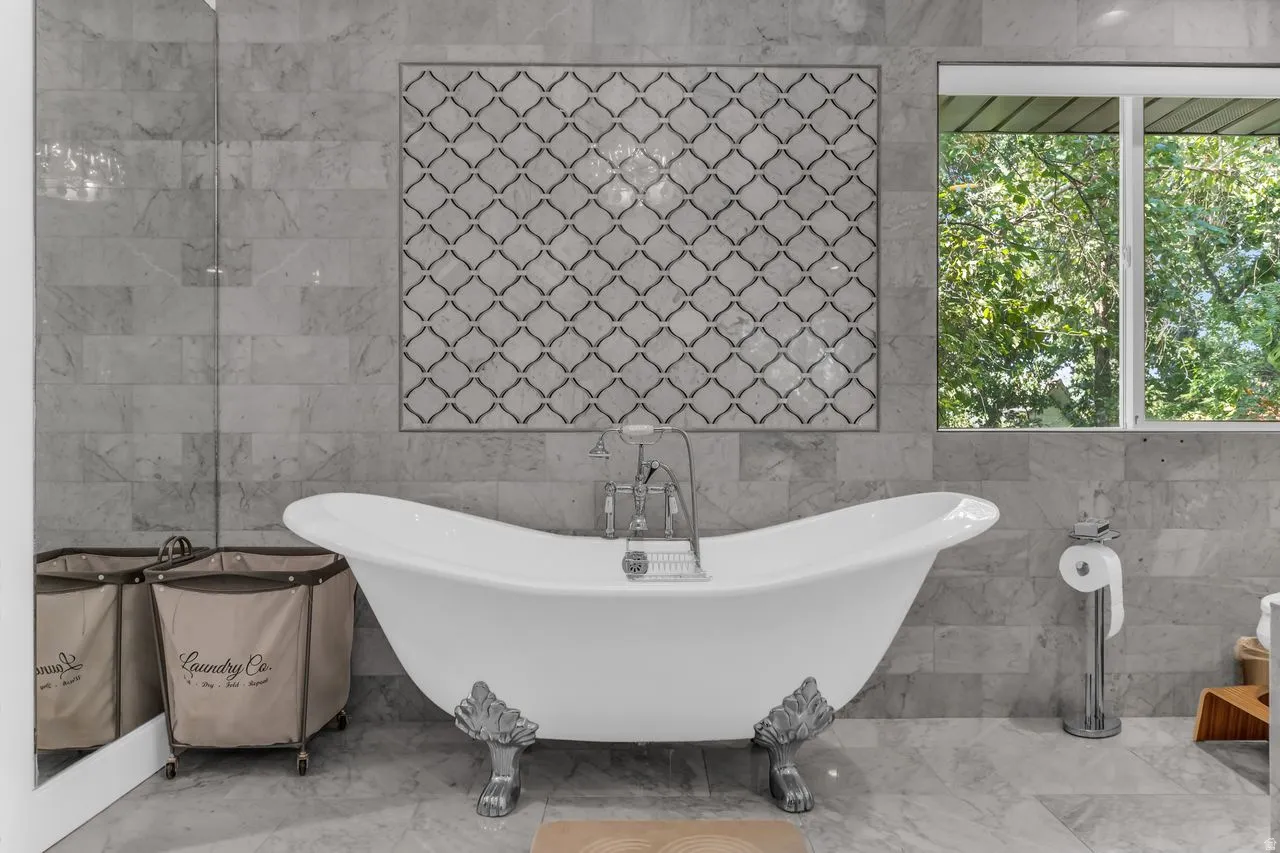 Freestanding clawfoot tub with polished chrome fixtures, set against large format grey tile walls and a large window