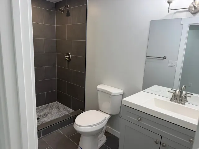 Bathroom featuring a walk-in shower with dark gray tile surround and mosaic floor, a toilet, a single vanity with a white countertop and undermount sink, and a framed mirror