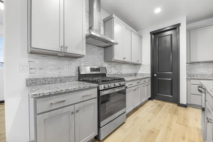 Kitchen featuring light gray cabinetry, stainless steel range with overhead ventilation, granite countertops, patterned tile backsplash, and light wood-finish flooring