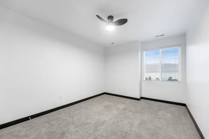 Spacious room featuring light gray carpet flooring, white walls, black baseboards, a ceiling fan with integrated lighting, and a window with horizontal blinds