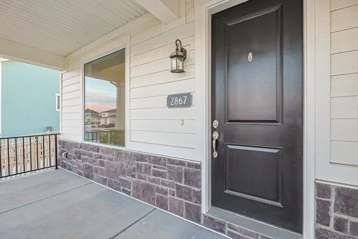 Front entry featuring a paneled black door with brass hardware, a large window, a stone veneer accent, white horizontal siding, and a covered porch with a metal railing