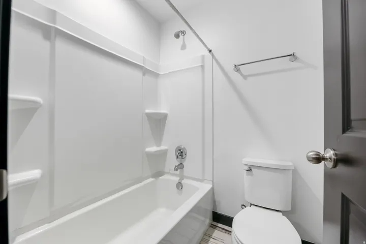 Bathroom featuring a white tub-shower combination with integrated shelving, chrome shower fixtures, a white toilet, and striped floor tiling