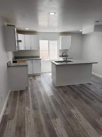 Kitchen featuring white shaker cabinetry, speckled countertops, a center island with a built-in sink and gooseneck faucet, recessed lighting, and wood-finish flooring
