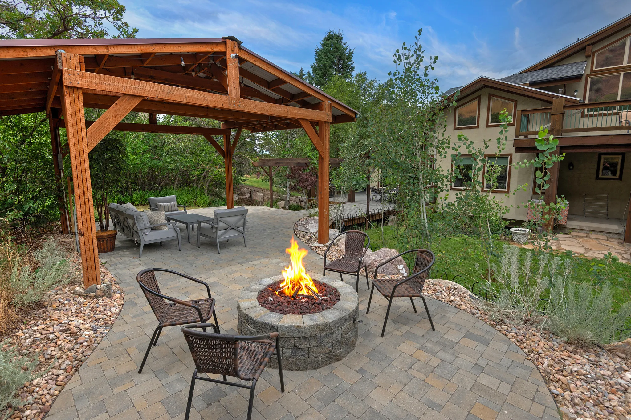 Expansive outdoor living area featuring a paver patio with a stone fire pit, a wood-frame covered pergola, and surrounding landscaping