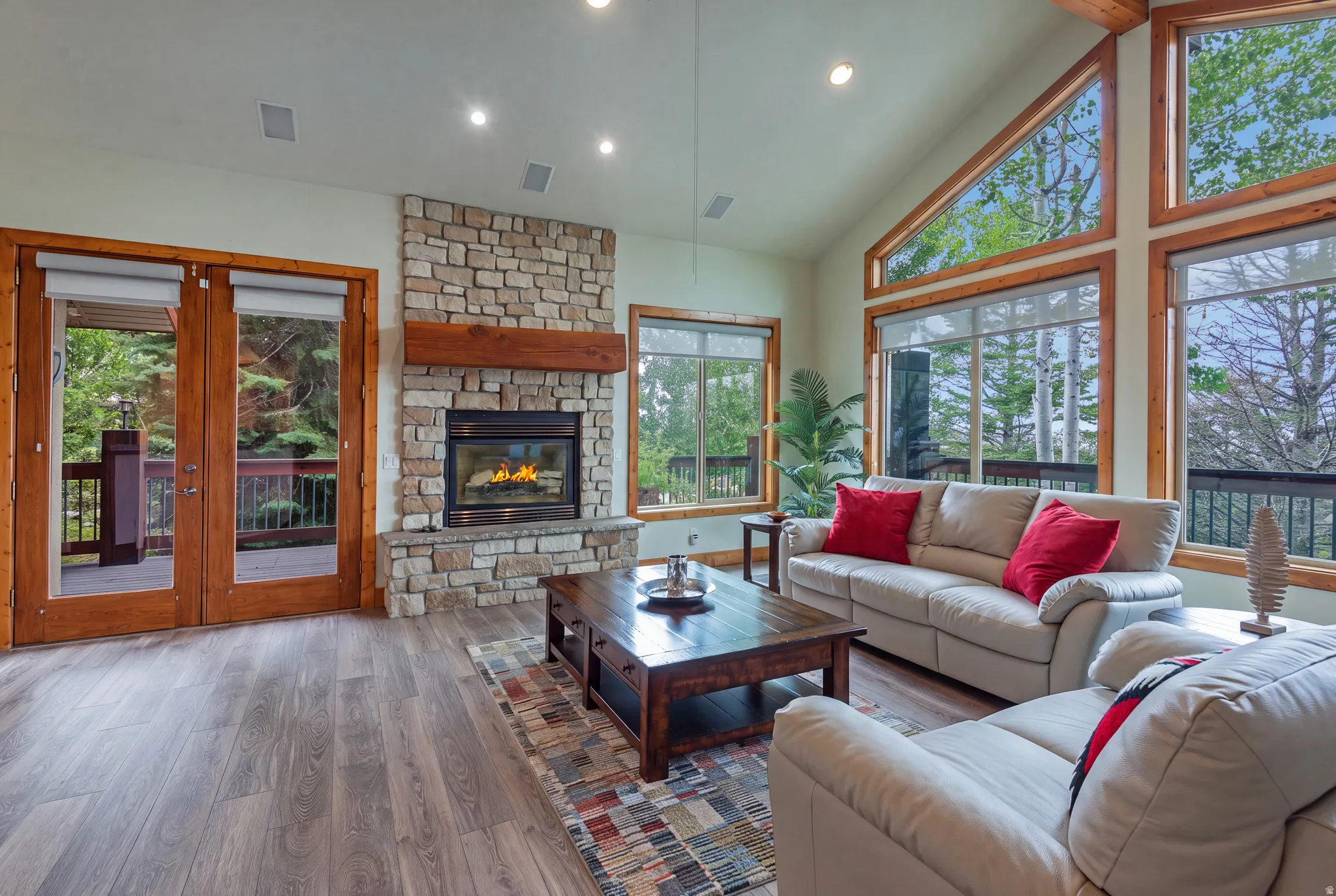 Vaulted ceiling living area featuring a stone fireplace with wood mantle, wood-finish flooring, recessed lighting, and large windows with integrated blinds