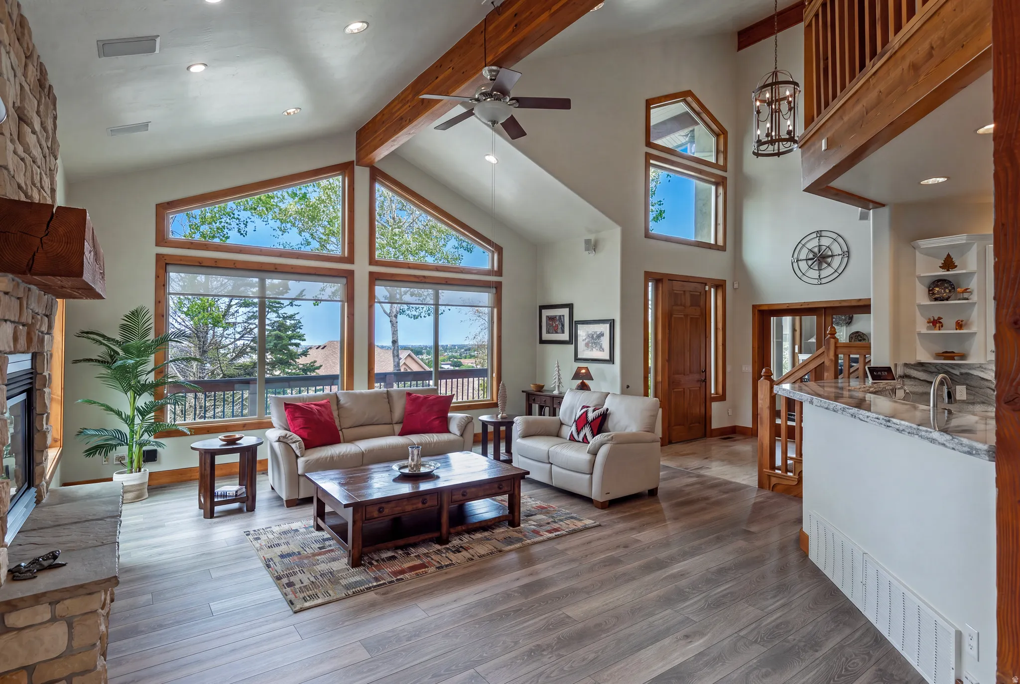 Vaulted ceiling great room featuring expansive windows, a stone fireplace, wood-finish flooring, exposed wood beams, and a wood-railed staircase
