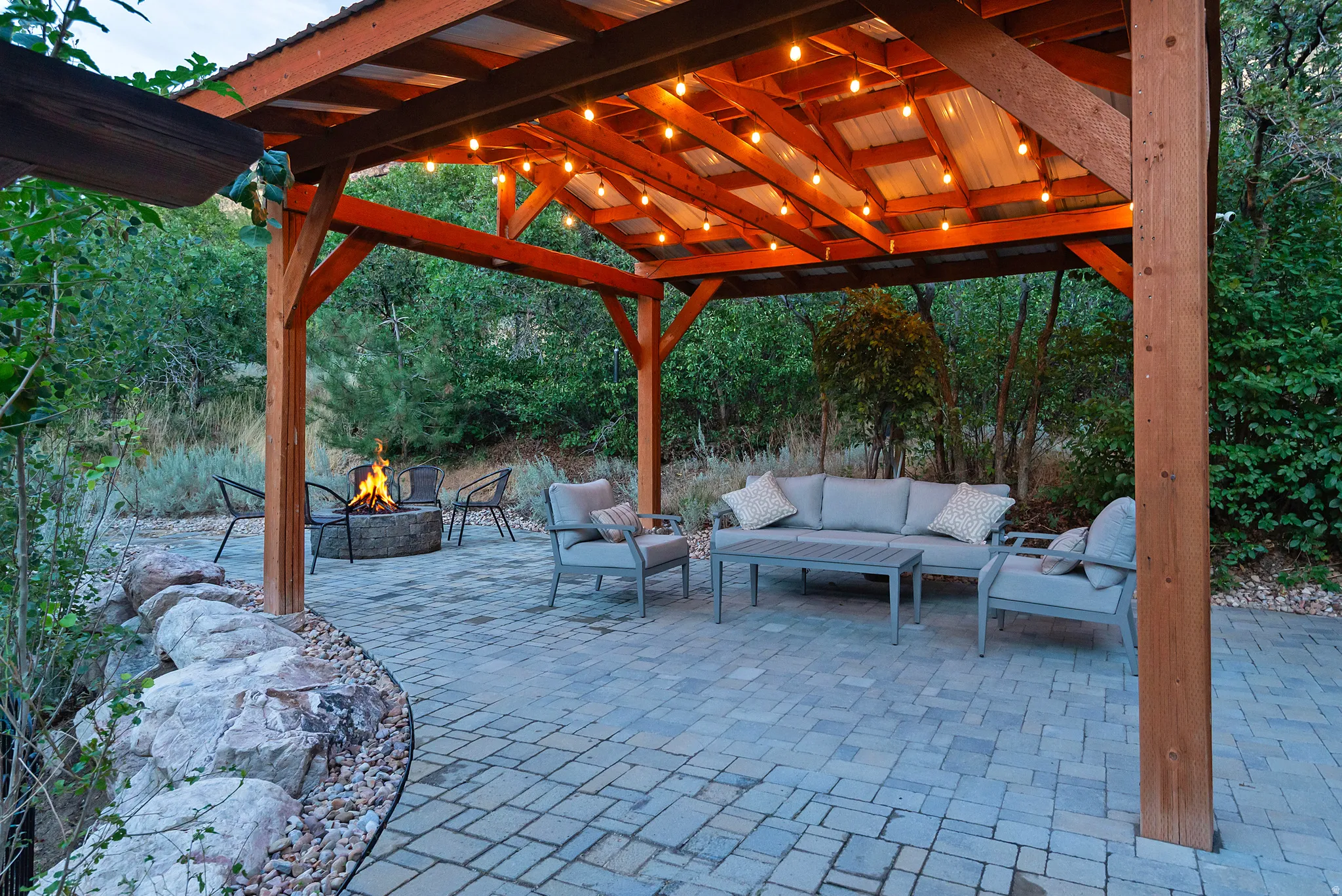 Expansive outdoor living area featuring a wooden pergola with string lighting, a stone fire pit, and a paver patio