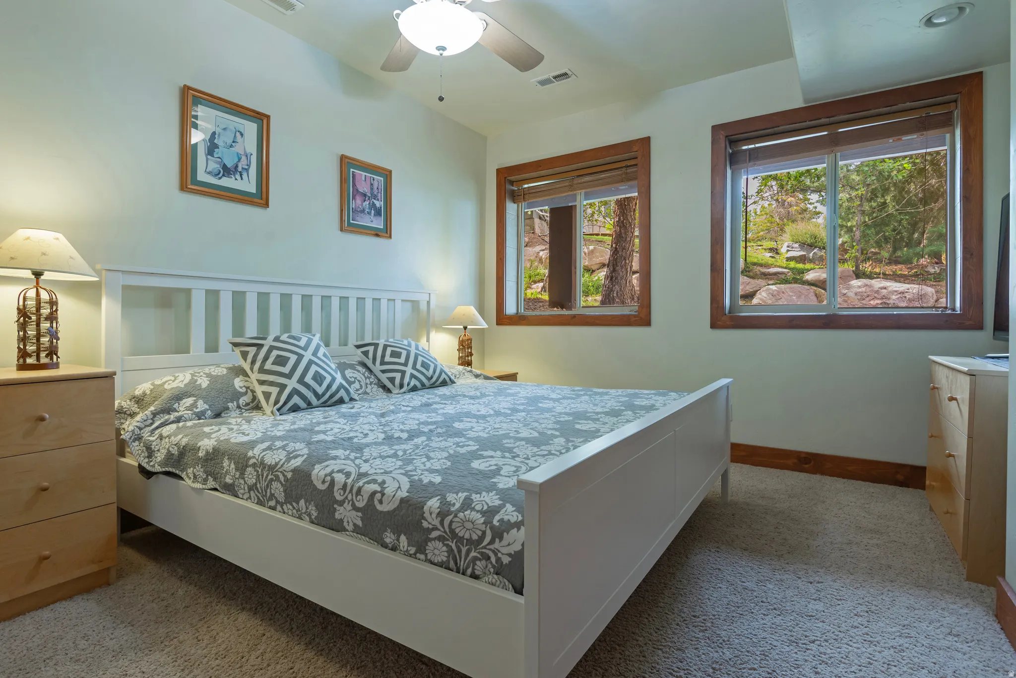 Carpeted bedroom featuring two windows with wood-trim framing, a white slatted headboard, and a ceiling fan with integrated lighting