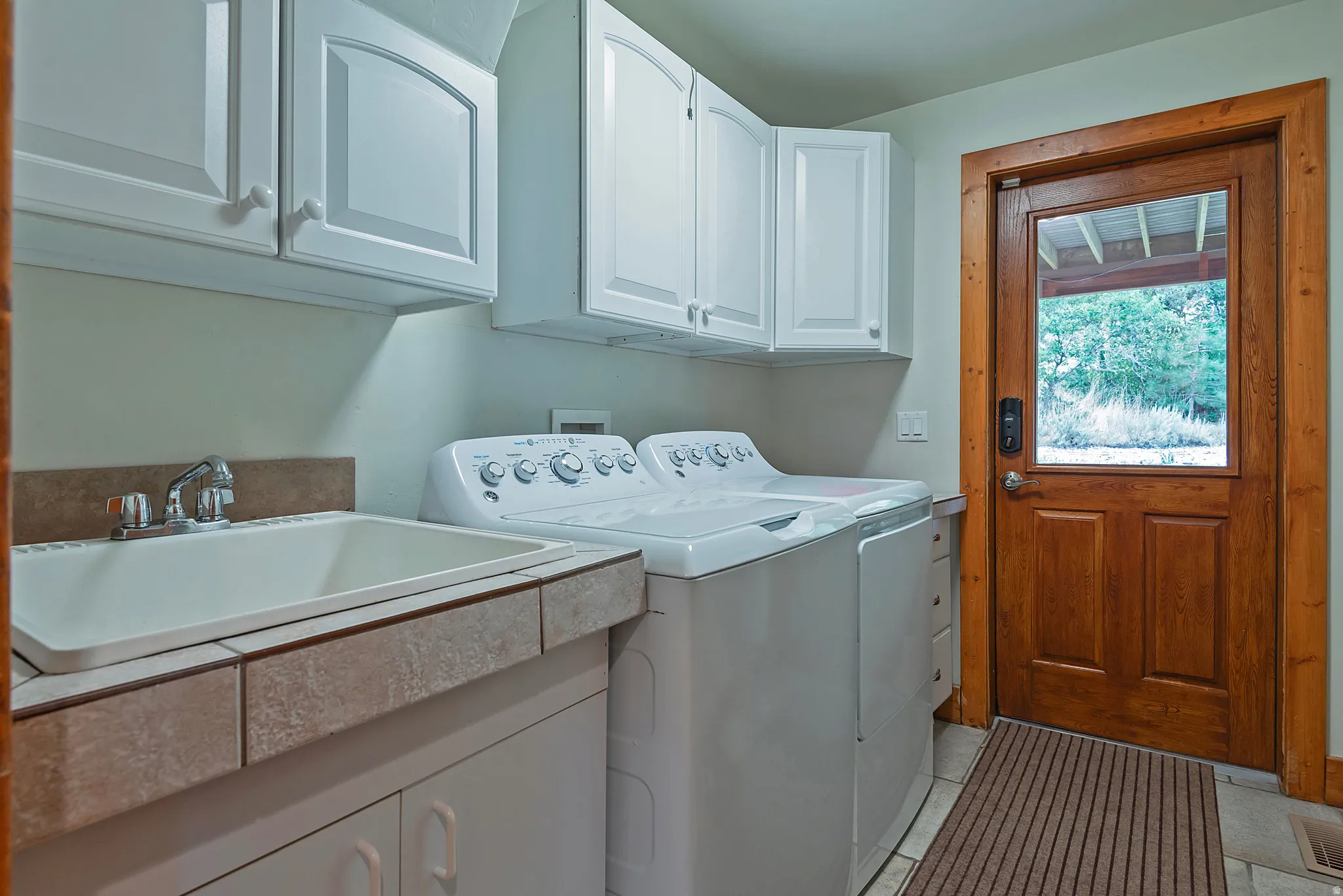 Utility room featuring white cabinetry, a built-in sink with tiled countertop, and wood-finish door with glass pane