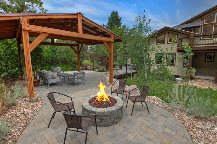 Expansive outdoor living area featuring a paver patio with a stone fire pit, a wood-frame covered pergola, and surrounding landscaping