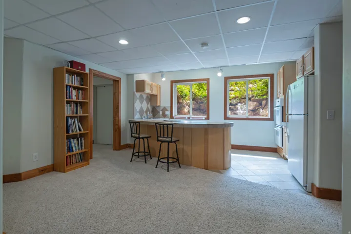 Lower-level recreation room featuring a built-in wet bar with wood cabinetry, two windows, recessed lighting, and a combination of carpet and tile flooring