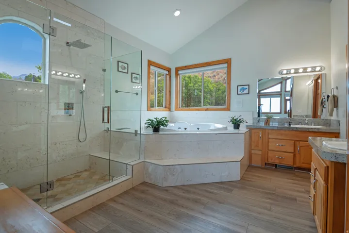Bathroom featuring a glass-enclosed shower with tiled walls and floor, a built-in jetted tub, dual vanity with wood cabinetry and stone countertops, and wood-finish flooring