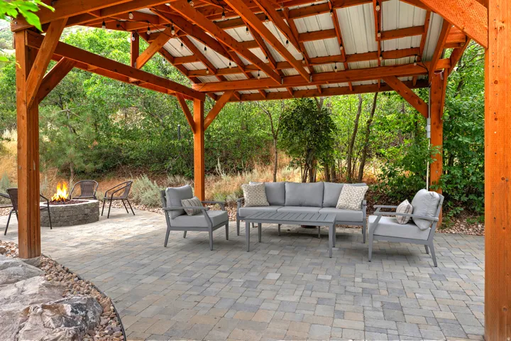 Expansive covered patio featuring a wood beam pergola with a metal roof, paver stone flooring, and a built-in stone fire pit