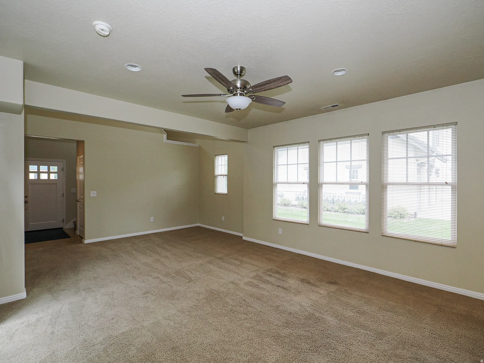 Spacious living area featuring neutral carpeting, three large windows with blinds, a ceiling fan with integrated lighting, and recessed ceiling lights