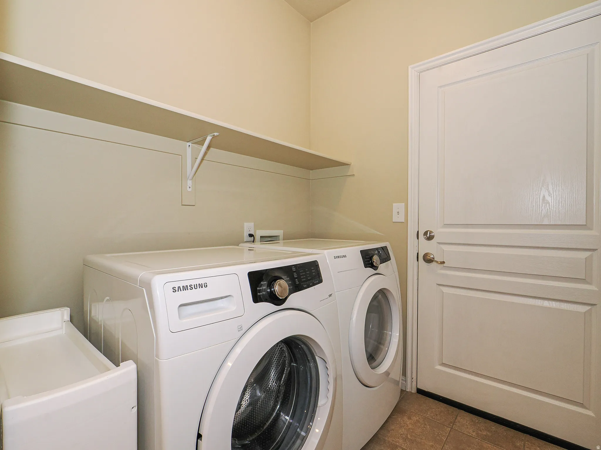 Dedicated laundry area featuring a white paneled door with brushed nickel hardware, a built-in wall shelf, light neutral wall paint, and tile flooring