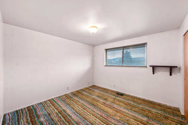 Room featuring a multi-colored striped carpet, a single window with horizontal blinds, a ceiling-mounted light fixture, and a wall-mounted wooden shelf