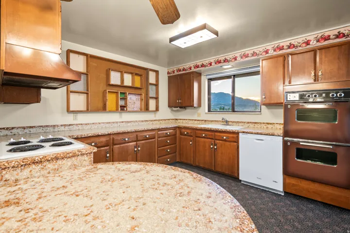 Kitchen featuring wood cabinetry, integrated double wall ovens, a cooktop with a coordinating range hood, a window over the sink, and speckled countertops
