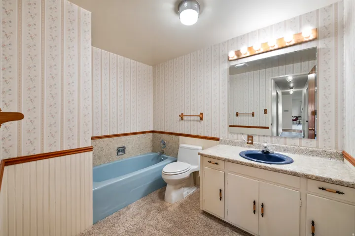 Full bathroom featuring a single vanity with a blue basin, a large wall-mounted mirror, a tub-shower combination, and wallpapered walls with beadboard wainscoting