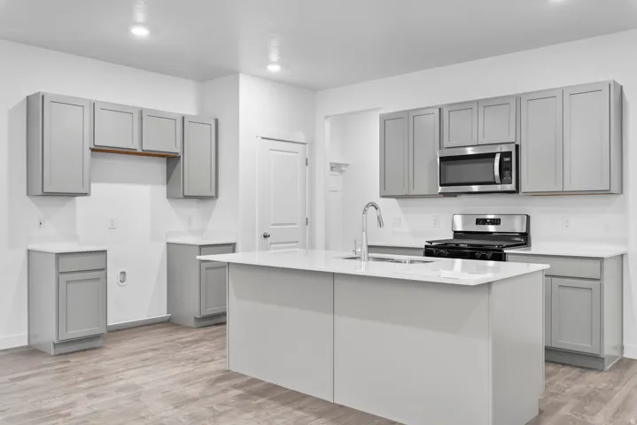 Modern kitchen featuring a central island with an integrated sink, light gray shaker cabinetry, quartz countertops, a stainless steel microwave, and a stainless steel gas range