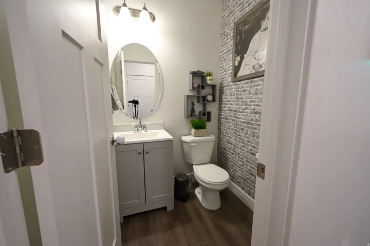 Powder room featuring wood-finish flooring, a contemporary vanity with an integrated sink, an oval wall-mounted mirror, and a decorative brick-patterned accent wall