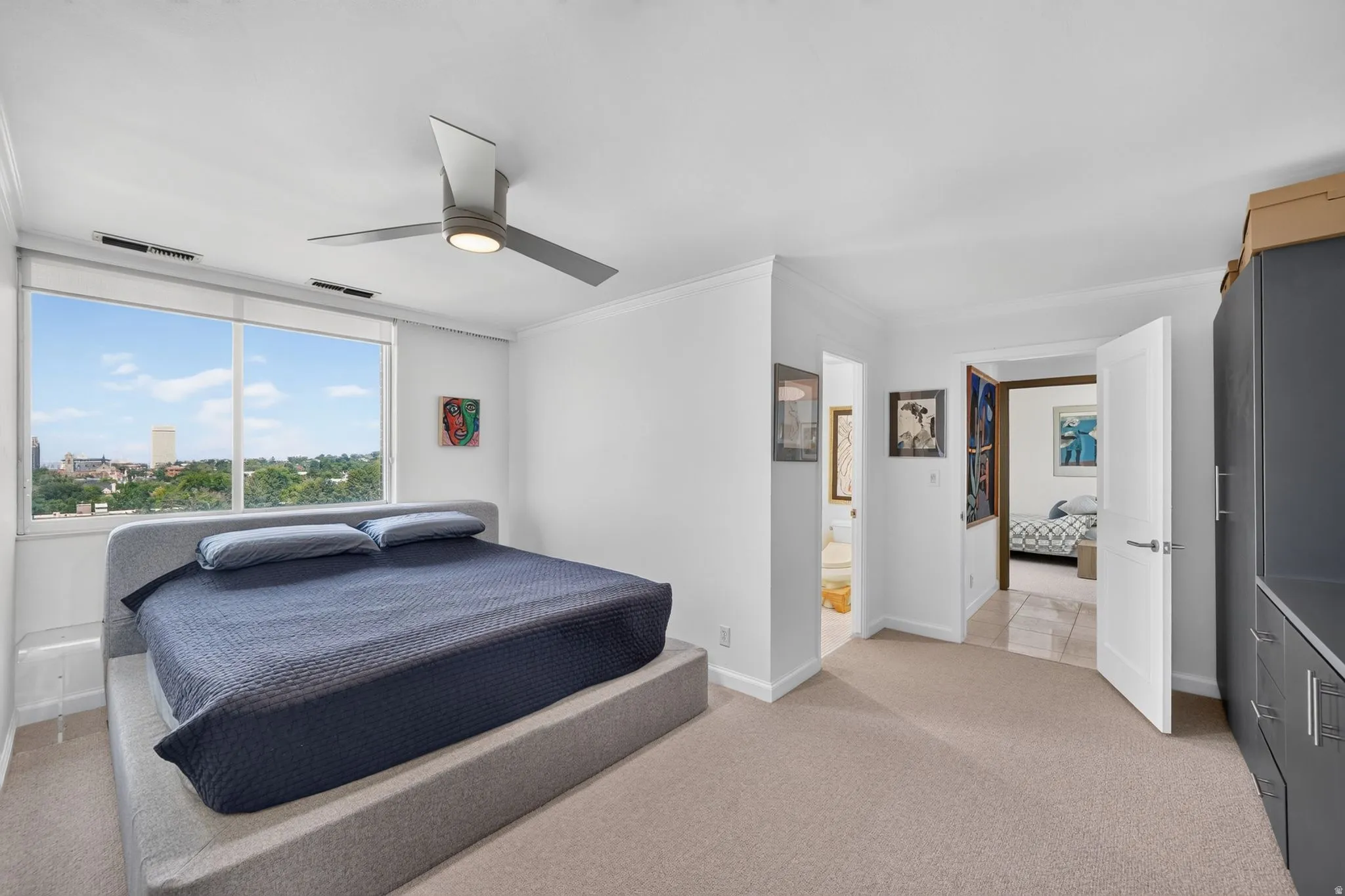 Window wall offering city and treetop scenery, carpeted flooring, contemporary ceiling fan with integrated lighting, white interior doors, and built-in gray cabinetry