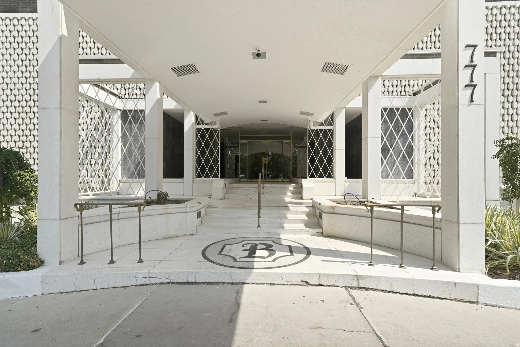 Covered entryway featuring decorative white lattice panels, a prominent B-initial floor inlay, built-in planters with decorative metal railings, a textured building facade, and large exterior numbers