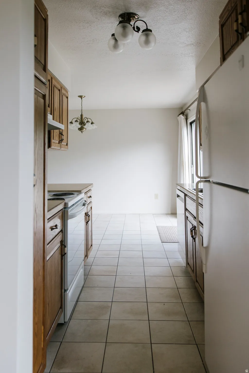 Galley kitchen featuring wood cabinetry, tile flooring, a built-in electric range, and a window with white curtains